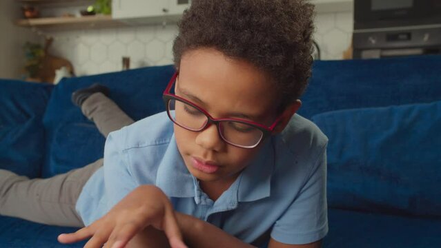 Closeup portrait of focused smart lovely African schoolboy in eyeglasses lying on couch, doing homework, learning and reading book while spending leisure in home interior.