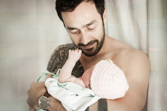 Young strong bare chested father with tattoo covering his shoulder and arm holds his newborn baby daughter wearing a pink wooly hat and smiles as he looks at her in a room in Edinburgh, Scotland, UK