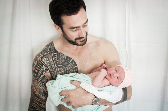 Strong bare chested father with tattoo covering his shoulder and arm holds his newborn baby daughter wearing a pink wooly hat and smiles as he looks at her in a room in Edinburgh, Scotland, UK