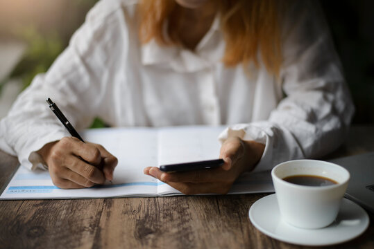 Planner using phone and write appointment to plan daily agenda calendar book. Woman Hand mark and note schedule on diary at business office desk. Coffee place on table. Work online at home concept.