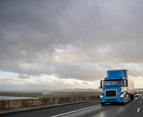 Fototapeta na wymiar Blue big rig semi truck transporting cargo in dry van semi trailer running on the wet highway road with stormy rain sky