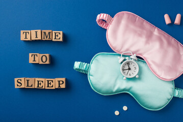Top view photo of the small white alarm on the two silk masks pink and light blue one with two pills wooden cubes with letters and two pink earplugs on the deep blue background