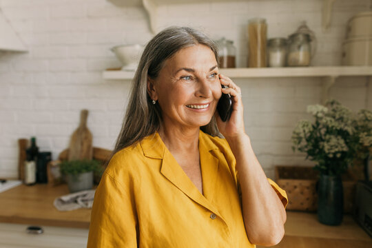 Close-up portrait of charming retired lady in yellow shirt having nice phone conversation isolated over kitchen interior background, looking through window during talk on black smartphone