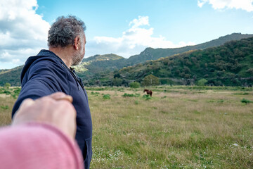 Fototapeta na wymiar Follow me. Man holds and pulls the photographer's hand while walking on the pasture and leads her towards the horses. Concept of union and new modern life in harmony with nature.
