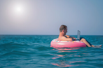 Close up male entrepreneur working on vacation with a laptop on inflatable ring in the water of sea or ocean. Copy space.