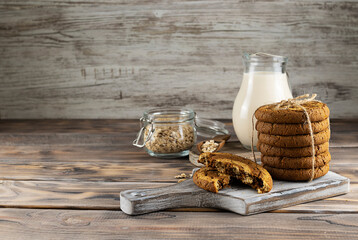 Oatmeal cookies are stacked on a light brown wooden board. Milk in a clear jug and oatmeal in a...