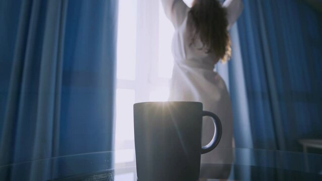 Woman in long bathrobe comes to open blue curtains on large window in spacious hotel room. Lady stretching hands after waking and take mug from glass table, selective focus on cup of coffee