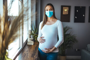 Young beautiful pregnant woman in medical surgical mask and white t-shirt quarantined at home by...