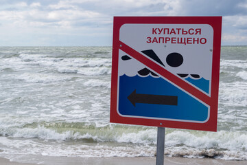 Sign with image of water, crossed-out swimmer, arrow and inscription in Russian IT IS FORBIDDEN TO SWIM. Plate installed on shore of sea, covered with foamy waves, with blurry cloudy sky on horizon.