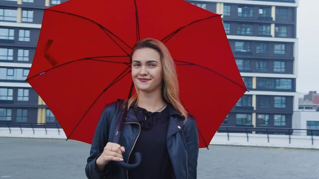 Cute lonely student with backpack is waiting for soul mate or boyfriend on street in rain. Attractive girl stands on roof under red umbrella. Beautiful model posing for photographer at photo shoot