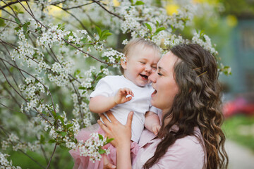 Lovely beautiful feminine mom with her daughter in her arms in spring with cherry blossoms. female natural beauty and beautiful wavy hair. Young European woman in a dress with a baby in a skirt.