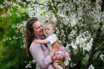 Tender mother in a pink dress holds a baby daughter in spring. Motherhood and femininity. Mom with long curly hair.