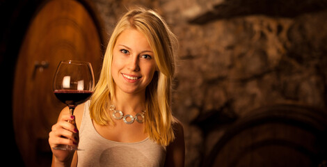 Beautiful young blond woman tasting red wine in a wine cellar - 484375019