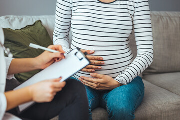 A pregnant Caucasian woman is indoors in a doctor's office. Her female doctor is wearing medical clothing. The woman is holding her stomach which the doctor writes on a clipboard.
