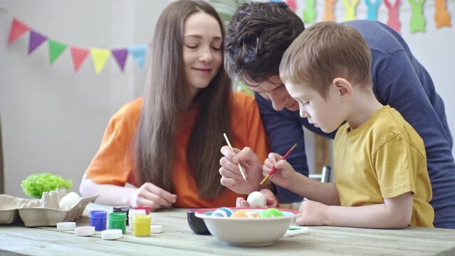 Mom, dad and little son in bright clothes are coloring eggs in a decorated room. Concept of family preparation for Easter, festive spring mood