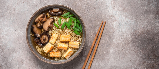 Asian vegan ramen noodle soup with roasted tofu cheese, chives and shiitake mushrooms in a bowl on brown grunge background. Top view. Selective Focus. Banner - obrazy, fototapety, plakaty