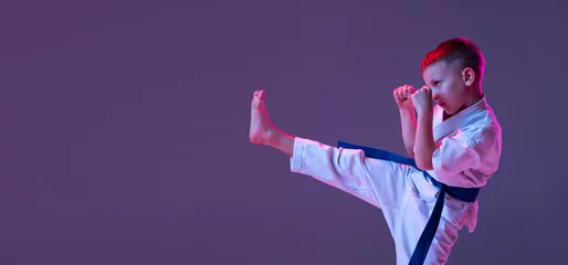 Afwasbaar fotobehang Portrait of sportive kid, male taekwondo, karate athletes in doboks doing basic movements isolated on purple background in neon. Concept of sport, martial arts © master1305