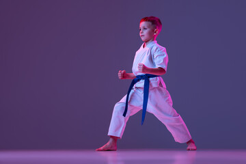 Dynamic portrait of kid, young male taekwondo, karate athletes in doboks doing basic movements isolated on purple background in neon. Concept of sport, martial arts © master1305