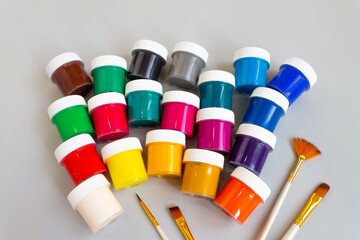 A set of closed jars with gouache of different colors and a brush for drawing on a gray background....