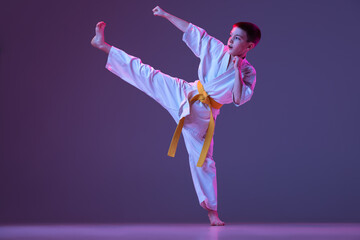 Dynamic portrait of kid, young male taekwondo, karate athletes in doboks doing basic movements isolated on purple background in neon. Concept of sport, martial arts © master1305