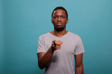 Portrait of young man pointing index finger at camera, standing over blue background. Focused person indicating direction and making choice to choose someone in studio. Individuality pick