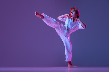 Portrait of sportive kid, male taekwondo, karate athletes in doboks doing basic movements isolated on purple background in neon. Concept of sport, martial arts © master1305