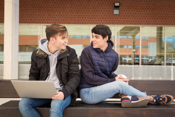 couple of two gay university students sitting on a bench with laptop and mobile phone looking at...