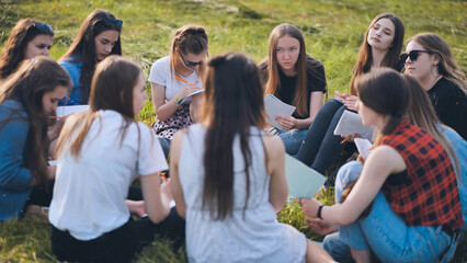 A group of female students are sitting in a circle on a meadow for collective work with notebooks.