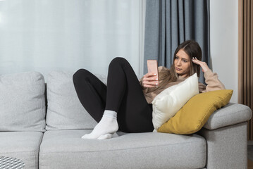 Obraz premium Smiling teenager girl relax lying on sofa at home using cellphone texting chatting with friend, happy young woman on couch hold smartphone shop online or check mobile application or social media