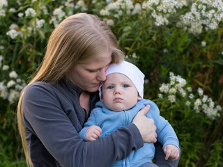 Medium horizontal portrait of pretty young blond mother cuddling baby boy with grumpy expression during late summer afternoon