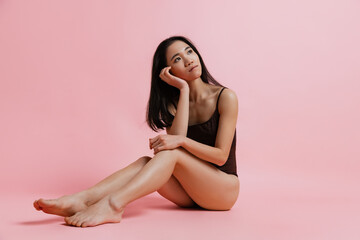 Obraz na płótnie Canvas Studio portrait of dreamy looking tender woman sitting and posing in bodysuit isolated over pink background