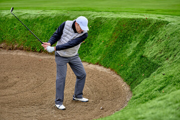 The golfer comes out of the bunker by hitting the ball with a golf club, on a winter day with wet...