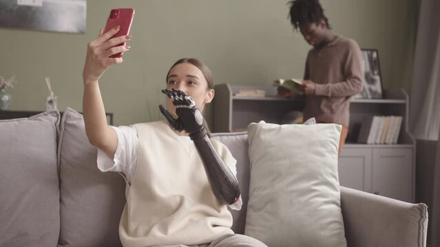 Medium slowmo shot of attractive young Caucasian woman with bionic arm taking selfie portrait on smartphone sitting on couch at home while her African-American boyfriend reading book in background