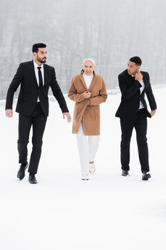 full length view of interracial bodyguards in black suits escorting mature businesswoman on snowy field.