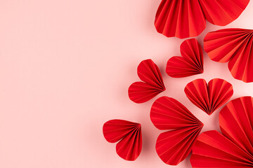 Bright red hearts of folded of chinese paper fans fly on pastel pink background as romantic Valentine day background, sideways border, top view, copy space.