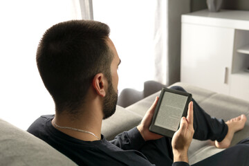 Unrecognizable young man reading with his e-book while sitting on the sofa at home.