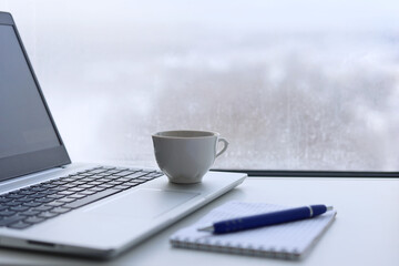 Coffee cup, laptop and notepad with pen on a table against the window, view to winter city. Cozy workplace in home office