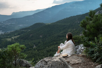 Naklejka na ściany i meble Young beautiful woman with long hair is sitting with her back, meditating and enjoying freedom with a beautiful view of the mountains and the forest. Travel and healthy lifestyle.