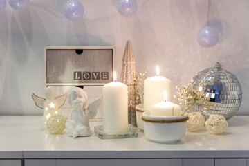 Fototapeta na wymiar The concept of decorating the house for Christmas. Lighted candles, a garland, a disco ball, an angel figurine on a white chest of drawers. Place for text