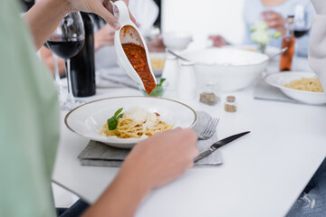 cropped view of woman pouring bolognese sauce in pasta with grated cheese on plate.