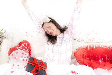 A beautiful woman in pink pajamas with a heart-shaped balloon lies on bed at home. Woman on Valentine's Day, birthday or anniversary. Love symbol