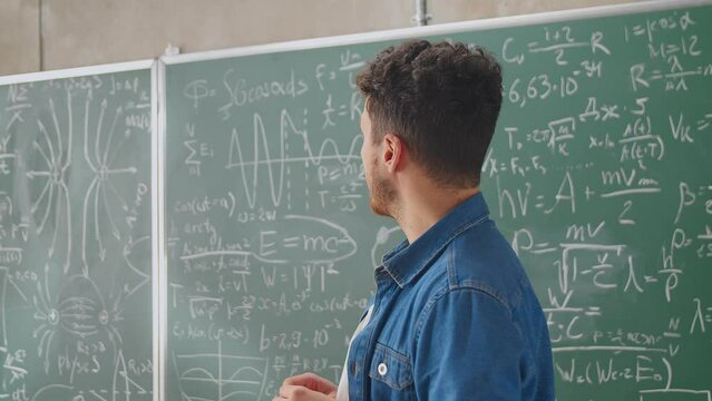 Caucasian young male teacher standing at blackboard with formulas, graphics and laws and explaining with finger. Lecturer of mathematics at school or college