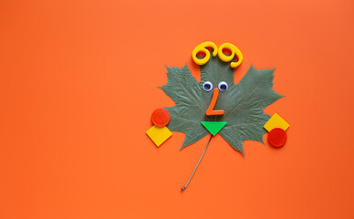 Orange background with dry marple leaf with funny face, geometric shapes and numbers.