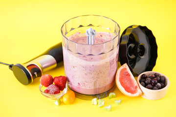 Black plastic electric hand blender and accessories with fruits and smoothie on yellow background.