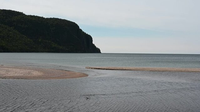 Water Swerves Around Two Sand Bars On Lake Superior With A Rock Face In The Background. 