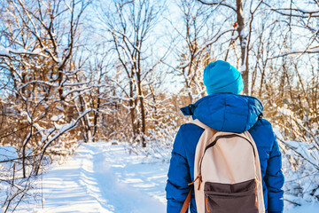 Rear view of a young man in a hat and with a backpack walking along a snowy path in the forest on a sunny day in winter
