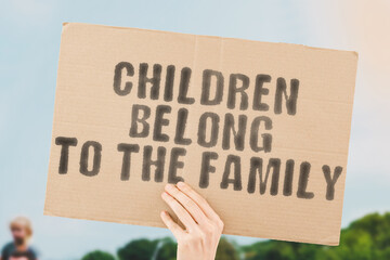 The phrase " Children belong to the family " on a banner in men's hand with blurred sea on the background. Bulling. Assistance. Afraid. Age. Care. Trouble. Poverty. Worry. Son. Suffer. Relation