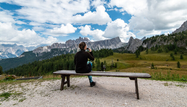 Young woman sitting at bench, drinking glass of red wine, looking at Cima Ambrizzola in front of Rifugio Cinque Torri. Dolomites, Trentino Alto Adige region, South Tyrol, Italy, Europe.