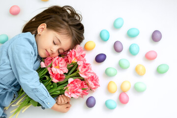 Obraz na płótnie Canvas A sweet girl with her eyes closed tenderly embraces a bouquet of tulips. Easter colored eggs are lying next to the child