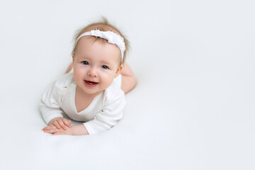 A little six-month-old girl in a white bodysuit and a headband is smiling sweetly lying on her tummy. Happy healthy baby